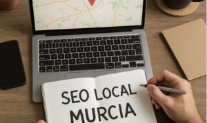 Diseño Web en Murcia Una persona escribe "SEO LOCAL MURCIA" en un cuaderno sobre un escritorio con un ordenador portátil que muestra un mapa, un bolígrafo y un cuaderno cerrado.